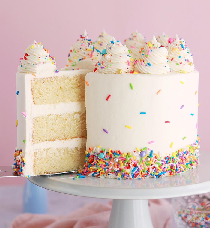 A vanilla cake with a slice cut out of it covered in frosting and sprinkles at the top and bottom.